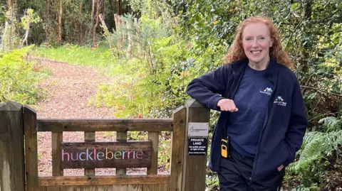 Julia Moore/ BBC A woman, Beverley Cook, stood next to a wooden gate in a forest. The gate has a multi-coloured sign on it reading "huckleberries"