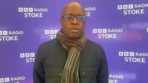 A man in glasses, wearing a scarf over a jacket is looking at the camera. He is standing in front of purple backdrop with BBC Radio Stoke branding on it