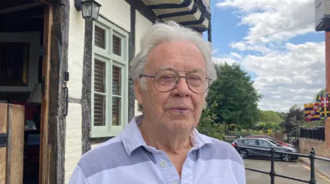 A photo of the chair of Tewkesbury Civic Society's Alan Purkiss. He has white hair and is wearing glasses and a light blue top. He is standing outside a building with a number of parked cars in the background.