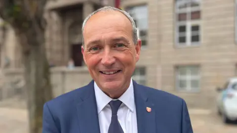 A man with short grey hair, wearing a navy blue suit, white shirt and blue tie. He is staring directly at the camera and smiling. The Royal Court is visible behind him.