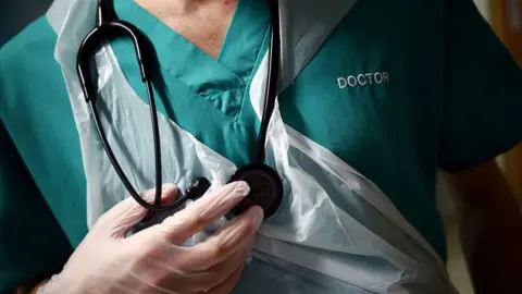 A close-up photograph of a healthcare worker’s torso wearing green surgical scrubs. The word “DOCTOR” is embroidered in white on the upper chest. A black stethoscope hangs around the neck, and one hand, covered by a light-coloured medical glove, is holding the stethoscope’s chest piece. A transparent disposable plastic apron is worn over the scrubs. 