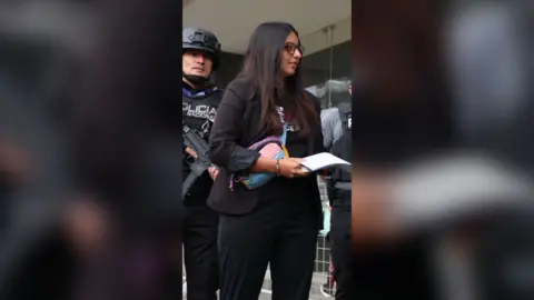 A woman with long dark hair wearing glasses and dressed in black, holds a piece of white paper