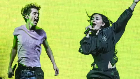 Getty Images Troye Sivan and Charli XCX on stage. On the left stands Troye, a man wearing a pink T-shirt, jumping up while Charli, in black, sings into a microphone with her left arm outstretched. Behind it is a lime green electronic backdrop.