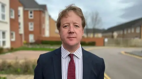Paul, a man with small brown hair. He is wearing a white and blue striped shirt and red tie and a black blazer. He is standing in the street.