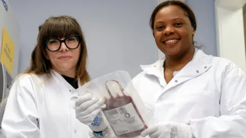 Mina Stoddart-Stones (right) is standing next to Joanne Mathews, both wearing white coats. Mina is holding a bag of her blood. They are both smiling.