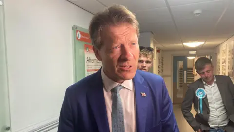 Richard Tice, the deputy leader of Reform UK, talks to members of the media. He is wearing a blue suit with a union flag pin badge on the lapel, a pink shirt and grey-blue tie. He is standing in a white coloured corridor with two men in suits standing nearby.
