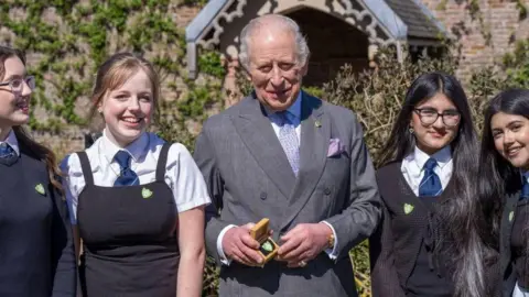 King Charles, wearing a grey suit holding a small box with a green badge inside, he is standing beside four school girls in uniform.