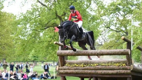 Badminton Horse Trials David Doel in a red riding coat leaps with Galileo off a very large bridge which is broken in half on the cross country course in 2022.