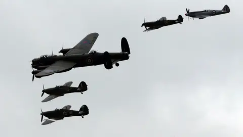 Four aeroplanes are silhouetted against a grey sky. The largest is the Lancaster bomber, and alongside it, are four spitfires.