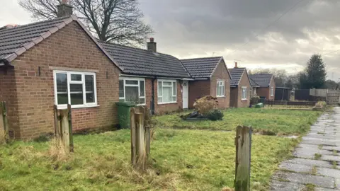 A row of bungalows. There is a fence missing and the front lawns appear to be overgrown.