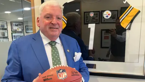 Feargal McCormack is holding an NFL ball. He's wearing a navy suit with a white handkerchief in his pocket and has a green spotty tie on. On the wall behind him is an NFL jersey saying number one in a frame. 