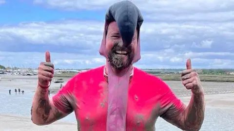 Jim Rugg Jim Rugg with a big smile on his face and giving a double thumbs up. He is wearing a muddy pink flamingo outfit with a large black beak drooping over his face. Behind him is the muddy river through which he has just ran.