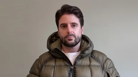Alex Grossomanides has brown hair, brown eyes and a beard. He is wearing an olive green puffer coat and looking at the camera while standing in front of a white wall indoors.