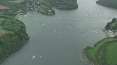 The image shows an aerial view of the Helford River estuary winding through the Cornish countryside, with wooded shorelines, patchwork fields and small settlements on either side. Dozens of boats can be seen moored across the water, while a motorboat leaves a visible wake as it travels along the river.