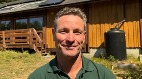 Greg Morel is standing outside a wooden building on a sunny day. He is wearing a dark green polo shirt. Behind him is a timber-clad building with large windows, a wooden deck and steps, and solar panels installed on the roof. A large black water tank stands beside the building, while dry grass and low vegetation cover the foreground.