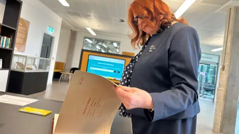 Anne flicks through an old record book with the title page register of books. She has shoulder length red hair and is wearing a smart navy suit.