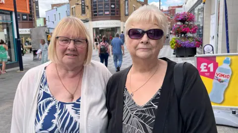 Phil Harrison/BBC A blonde woman in a white cardigan and blue and white top, wearing glasses. She is standing next to a woman, wearing sunglasses, and wearing a black cardigan and black and white top. They are standing at a high street.