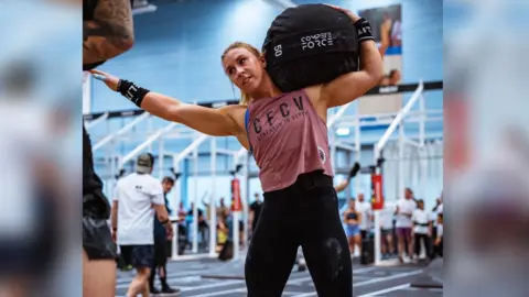 The picture shows Katherine Ogden in a red t-shirt and black leggings. She is training in a gym setting.
