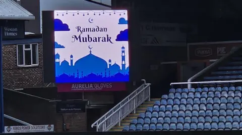 Shariqua Ahmed/BBC A white and blue billboard which says Ramadan Mubarak. It is in the corner of a football stadium, and empty seating is on the right of the picture.