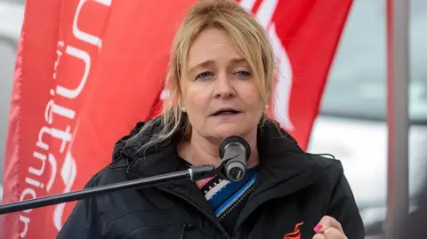 Getty Images A woman with long blonde hair and wearing a black coat talks into a microphone. A red Unite flag can be seen behind her.