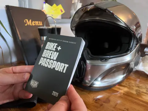A pair of hands holds a small book with the words "Bike and Brew Passport" on the front. There is a silver motorcycle helmet in the background. 