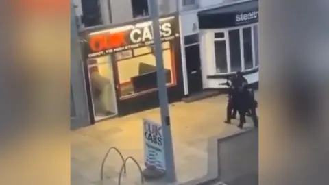 A still image of a video show three armed police officers approaching what looks like a taxi rank, with the sign Quik Cabs, where there is an individual in the doorway