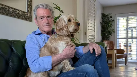 Jam Press Martin Dandridge in a blue shirt and jeans sat on a green sofa in a living-room with his light beige cockerpoo, Gulliver. 