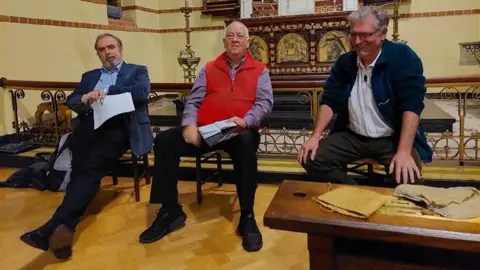 BBC Clockwise: Peter Hitchens and Richard Scrase sit in St Barnabus church, each on the side of debate moderator John Mair, during the e-bikes debate.