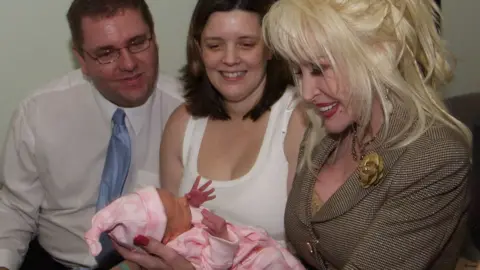 Three adults seated closely together indoors, focusing their attention on a newborn baby wrapped in a pink blanket. In the foreground, Dolly is holding the baby carefully in both hands. She is wearing a light brown checked suit jacket with decorative gold buttons and a flower-shaped brooch on the lapel. The baby is dressed in pink clothing and a matching hat, with one small hand raised upward. To the left of the image, Shane Jarman is seated wearing a white shirt, a light blue tie, and glasses. He appears to be looking down toward the baby. In the centre, Claire is seated wearing a white sleeveless top and is also looking toward the infant.