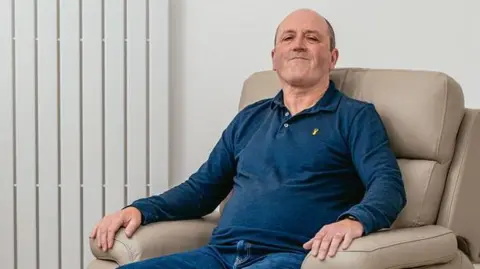 Owen Mathias/MNDA Mike Thomas, who is wearing a blue polo top, is sitting in a cream-coloured armchair and smiling while looking at the camera