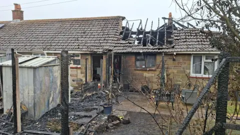 Bungalow after fire with charred remains of roof and blackened debris across a rear garden.
