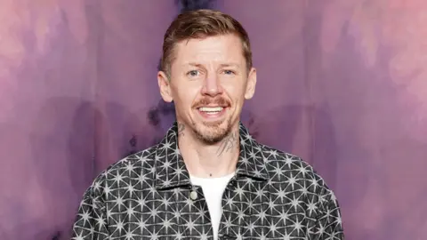 PA Media Professor Green, a middle-aged man with short gingery-brown hair, and a goatee and moustache, smiles at the camera. He has tattoos on his neck and is wearing a dark grey shirt jacket with a star pattern.