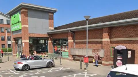Google The sunny outside of an Asda supermarket with customers leaving the entrance. A black clothes bank can be seen near to the entrance. An open top car is parked in a disabled spot near the doors and trolleys.