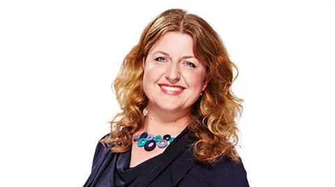 Global via PA Media A woman with shoulder-length curly hair smiles at the camera in a studio portrait against a plain white background. She is wearing a dark navy cowl-neck top and a chunky necklace made of blue and turquoise circular beads. The image is cropped from the waist up.
