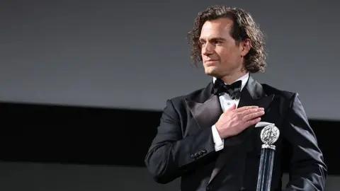 Getty Images Henry Cavill wears a suit and bowtie as he stands on-stage holding a tall, cylindrical award. He places his free hand flat across his chest and smiles appreciatively at someone off-stage.