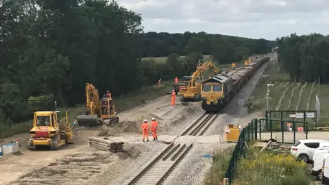 Construction work on east west rail line. Various yellow vehicles are working on the uninstalled lines, with construction workers dotted around the site wearing orang hi-vis jackets.