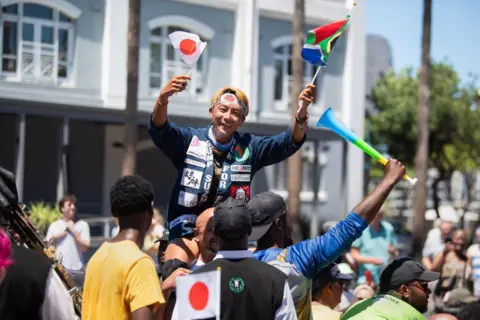 RODGER BOSCH / AFP A man waves the flags of Kenya and South Africa on Cape Town's waterfront. People hold him up in the air and celebrate with him.