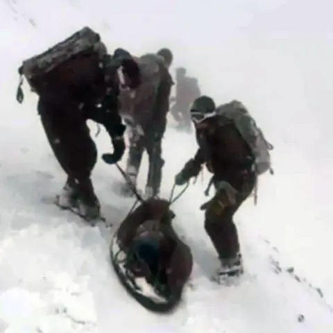 Royal Navy A group of marines dressed in green clothing during exercise on Ben Nevis. They are standing in snow and ice axes are laid on the ground. They are pulling the casualty in the makeshift stretcher.