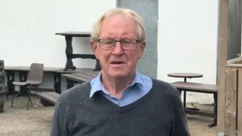 Limavady United Stanley Callaghan has short hair and is wearing glasses. He is wearing a blue shirt and grey anorak. 