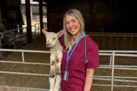 Karis Ramsay holding a lamb in front on an animal pen. Karis is wearing burgundy scrubs, a blue lanyard and ID as well and a stethoscope 
