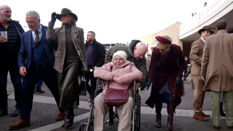 PA Media A woman in a wheelchair in a pink puffer jacket, white woolly hat and beige slacks is pushed by a man in a green jacket. He leans down to inspect the wheel, and is helped by Zara Tindall in a red headband, burgundy overcoat and black dress and boots. Other punters are visible behind them.