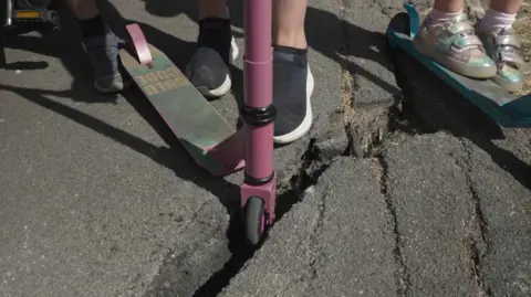 A mother demonstrates how easy it is to get a scooter wheel stuck in a crack in the pavement - this is close-up shot of the families' feet with a purple scooter's front wheel jammed.