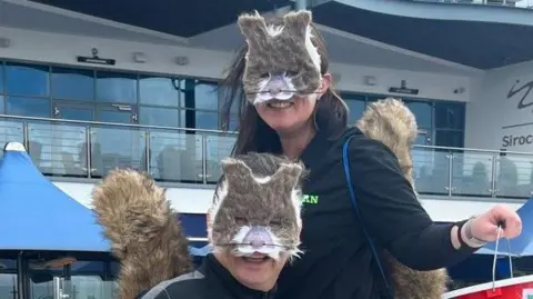 Dean and Kiley smile at the camera while wearing squirrel face masks and fluffy tails. They're also wearing black tops. Kiley is standing on a bench and is above dean and she's holding a fundraising bucket. 