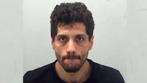 A police custody photo of Jan Drevenak, who has short black hair and a beard. He is wearing a black T-shirt and is standing in front of a grey wall.
