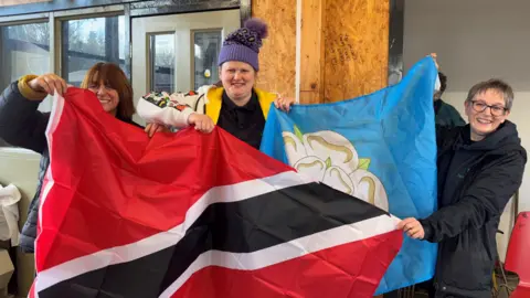 Three women indoors hold the Trinidad and Tobago flag and the Yorkshire flag, smiling for the camera.
