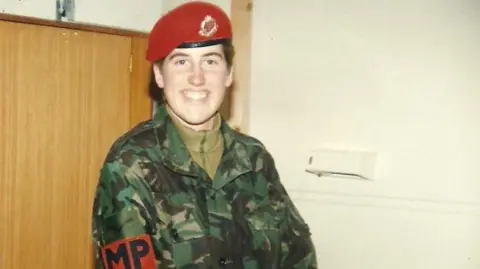 Kate Holvey-Williams Kate Holvey-Williams is smiling at the camera. She is wearing a red Military Police beret, a camouflage uniform and has a red armband with the letters MP. She is standing against a cream wall and wooden door.