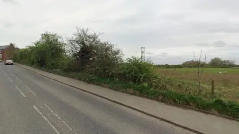 A street view taken on Philadelphia Lane. It is a single carriageway road with a pavement. In the distance is a red car and the start of a row of two-storey brick houses. Behind an unkempt hedge is a field with two white animals grazing.