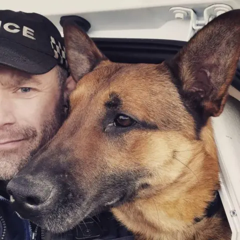 Humberside Police A Belgian Malinois dog who has a long nose, brown face and pointy ears is looking to the side. Next to him you can see half the face of a male police officer, who has brown stubble and moustache and is wearing a black police cap. They appear to be inside a vehicle.