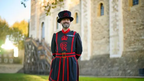 Historic Royal Palaces Ryan Brown with a short cropped beard smiling at the front of the tower of London. He is wearing a ceremonial blue and red tunic with a belt around its waist.  He is wearing a tall blue hat. The hat is trimmed with red. A Good Conduct medal for long service is pinned on the chest of his tunic which is a Yeoman Warder uniform. 
