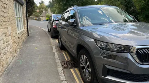 Two cars parked on double yellow lines on a road in the Cotswolds village of Bibury. One is a large grey Skoda and there is a smaller black car parked behind it.